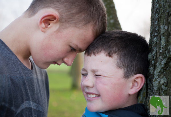 Bigger boy with a smaller boy backed up against a tree, head to head